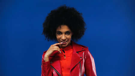 Smiling african american woman with finger near lips looking at camera isolated on blue