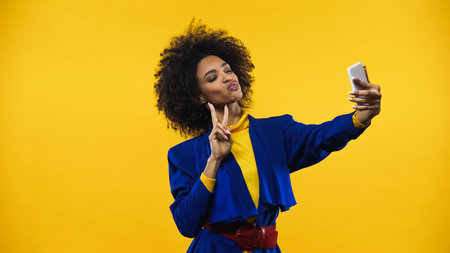 African american woman showing peace sign during selfie isolated on yellow