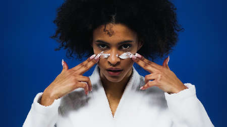 African american woman in bathrobe applying face cream isolated on blueの写真素材