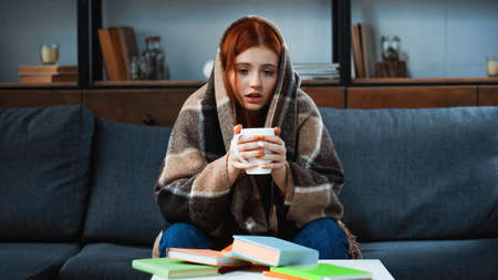 Girl in blanket holding cup near books on blurred foreground