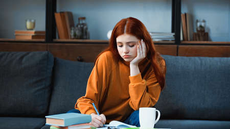 Upset student writing on notebook near cup and books