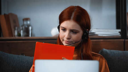 Student in headset looking at book near laptop on blurred foreground at home