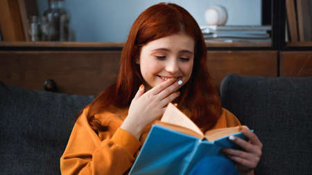Positive teenager with hand near mouth reading book on blurred foreground