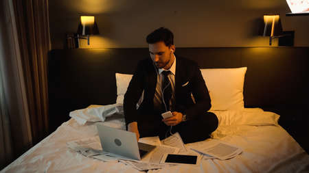 Businessman in earphones using laptop while holding smartphone near papers on hotel bedの写真素材
