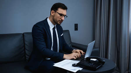 Businessman using laptop near papers and telephone in hotel room