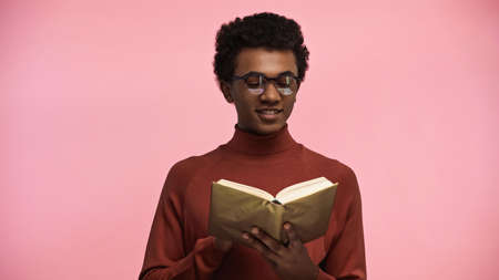 happy african american teenager in turtleneck sweater and glasses reading book isolated on pink