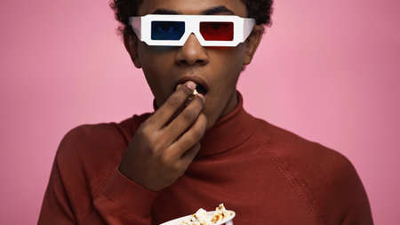 african american teenager in 3d glasses eating popcorn isolated on pink