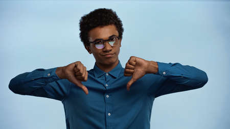 african american teenage boy in glasses showing thumbs down isolated on blue