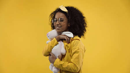 happy african american girl in glasses holding soft bunny isolated on yellowの写真素材