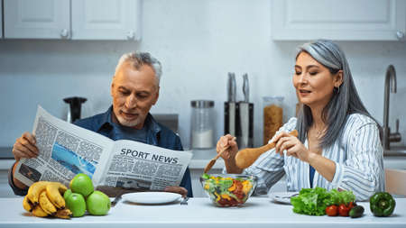 elderly asian woman mixing salad near husband reading sport news during breakfastの写真素材