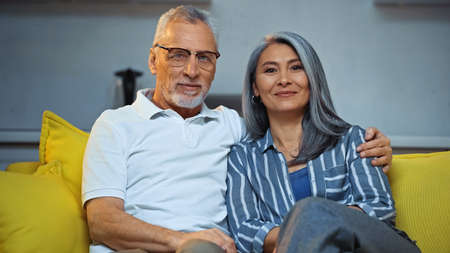 happy gray haired man embracing smiling asian wife while sitting on sofa at homeの写真素材