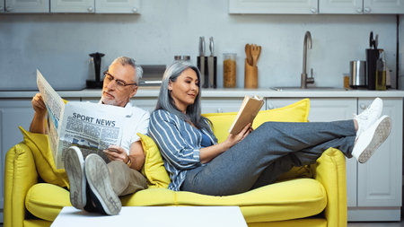 elderly interracial couple reading book and newspaper on sofa at homeの写真素材