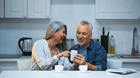 laughing asian woman pointing with finger at smartphone near smiling husbandの写真素材