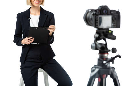 cropped view of smiling anchorwoman with clipboard sitting near digital camera on blurred foreground...