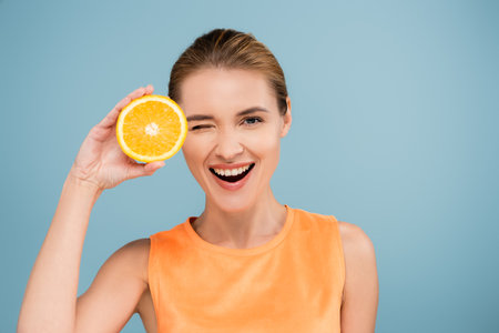excited woman winking at camera while holding half of ripe orange isolated on blueの写真素材