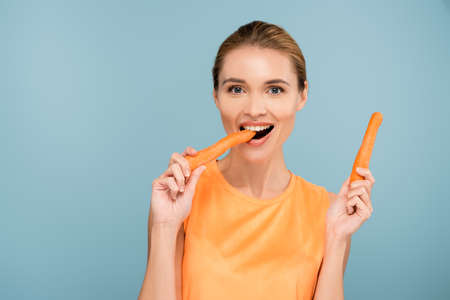 pleased woman looking at camera while eating whole carrot isolated on blueの写真素材