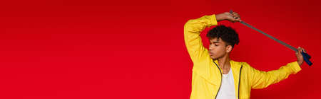 pierced african american man in yellow jacket posing with skipping rope isolated on red, bannerの写真素材