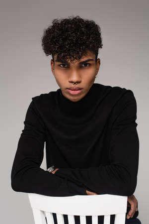 curly african american man in turtleneck sweater leaning on white chair isolated on gray