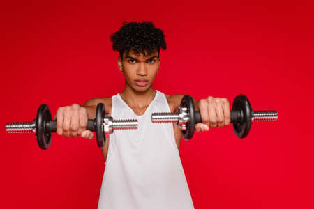 sportive african american man in tank top exercising with dumbbells on blurred foreground isolated on redの写真素材
