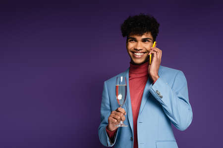 smiling african american man holding glass of champagne and talking on smartphone on purpleの写真素材