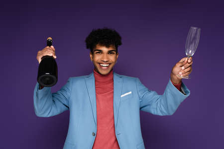 pierced african american man in blue blazer smiling while holding bottle of champagne on purpleの写真素材