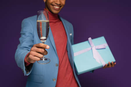 partial view of cheerful african american man holding glass of champagne and present on purpleの写真素材