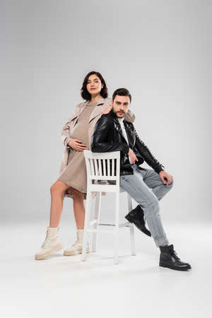 Pregnant woman looking at camera near stylish husband on chair on gray backgroundの写真素材