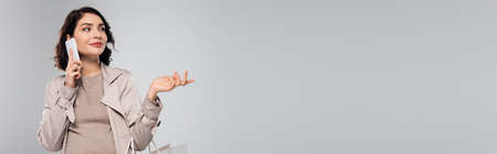 Pregnant woman talking on smartphone and holding shopping bag isolated on gray, banner