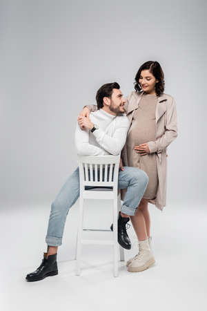 Smiling pregnant woman holding hand of husband on chair on gray backgroundの写真素材