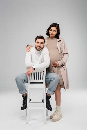 Pregnant woman hugging husband looking at camera on chair on gray backgroundの写真素材