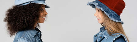 side view of smiling multicultural girls in stylish hats looking at each other isolated on gray,...