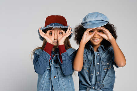 joyful multicultural friends in denim clothes and hats showing eyeglasses with hands isolated on grayの写真素材