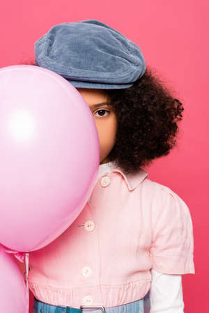 fashionable african american girl in stylish cap obscuring face with festive balloon isolated on...