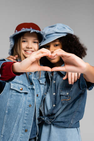 happy multicultural girls in stylish clothes showing heart symbol with hands on gray, blurred...