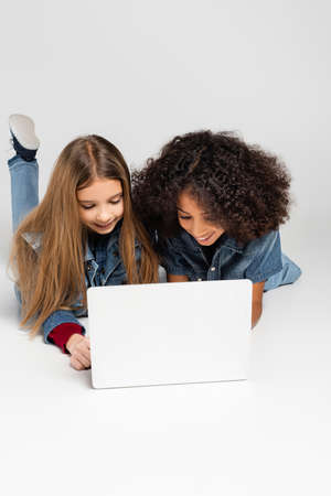 cheerful interracial friends in denim clothes using laptop while lying on gray