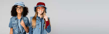 joyful interracial girls in trendy denim clothes, with paper cut eyeglasses, isolated on gray, bannerの写真素材