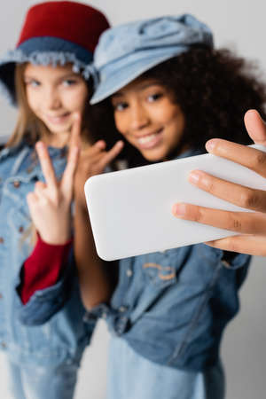 happy interracial girls showing victory and rock signs while taking selfie on gray, blurred...