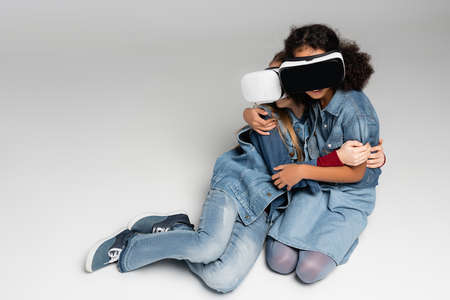 frightened interracial girls in vr headsets embracing while sitting on gray