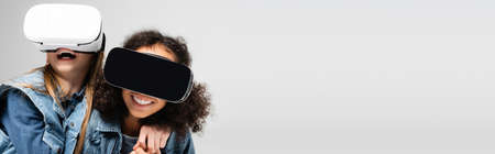 excited interracial girls having fun in vr headsets isolated on gray, banner