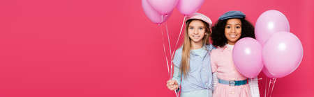 stylish interracial kids holding festive balloons while posing isolated on pink, bannerの写真素材
