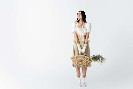 full length of joyful young woman in spring outfit holding straw bag with flowers on white