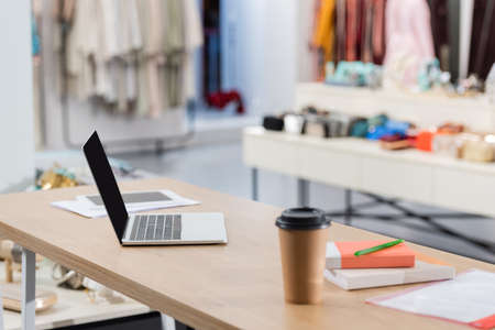 Laptop near notebooks and coffee to go on blurred foreground in showroom