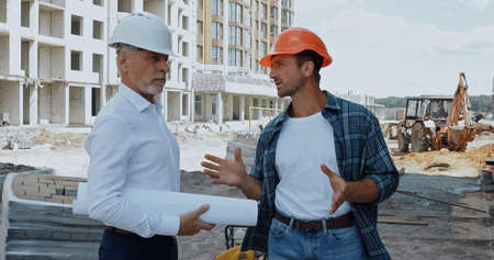 Engineer with blueprints talking with builder in hard hat on construction siteの写真素材