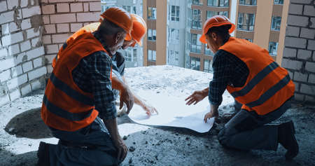 Three builders in hand hats discussing blueprint while sitting on construction site