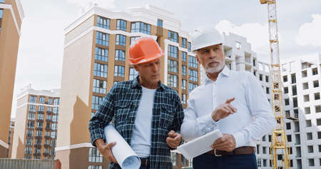 mature engineer and builder with gadget and blueprint talking on construction siteの写真素材