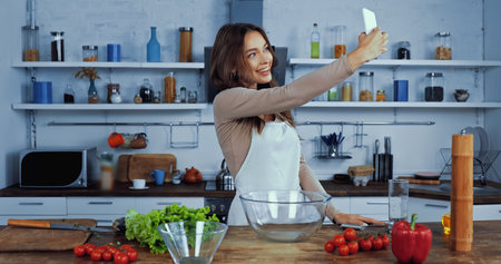 cheerful woman in apron taking selfie near ingredients on tableの写真素材
