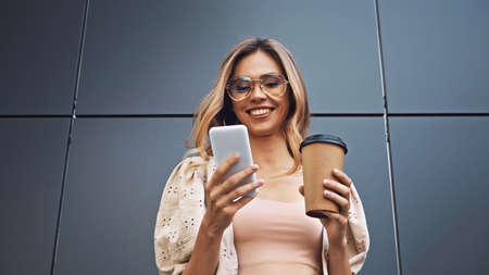 happy woman holding coffee to go and using smartphone near buildingの写真素材