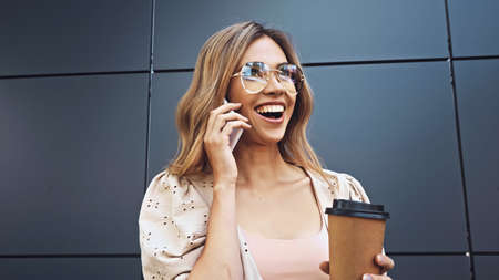 cheerful woman holding coffee to go and talking on smartphone near buildingの写真素材
