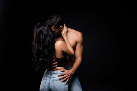 Shirtless man kissing brunette girlfriend in bra and jeans isolated on blackの写真素材
