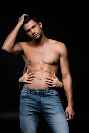 Sexy man looking at camera while girlfriend touching torso isolated on blackの写真素材
